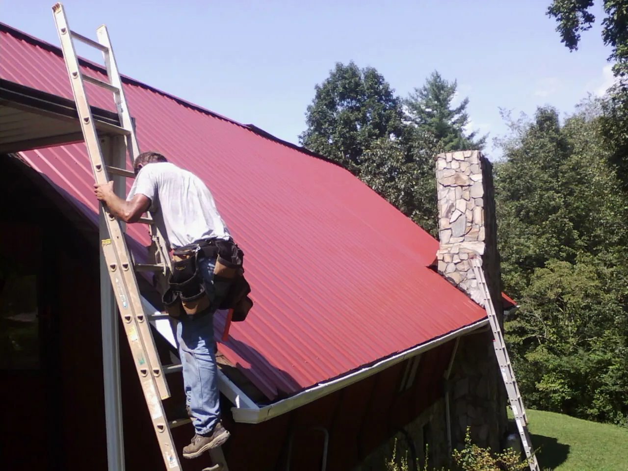 Roofer on a ladder beside a red metal roof and stone chimney — Soffit & Fascia Repair by Roofing Brothers in Frankewing